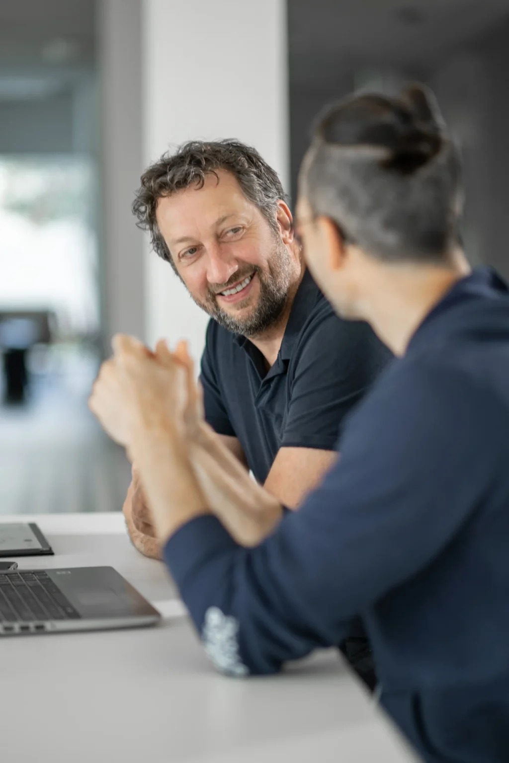 Man, smiling, wearing dark blue shirt, sitting with laptop at side of desk