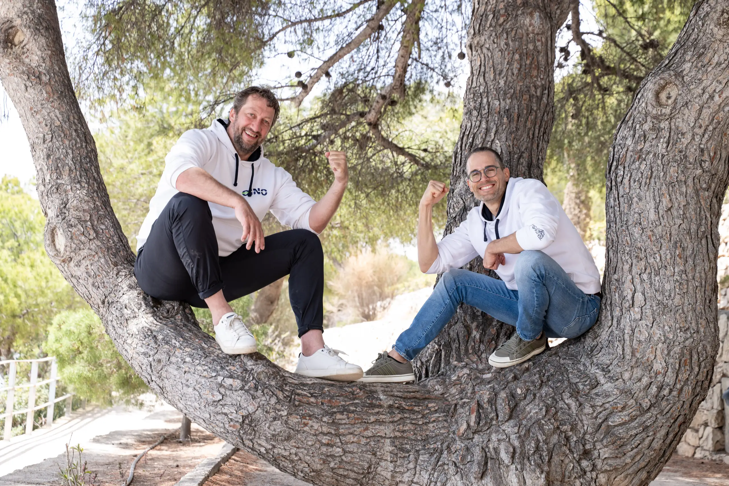 Two men in white hoodies pose laughing in a tree, showing their arms