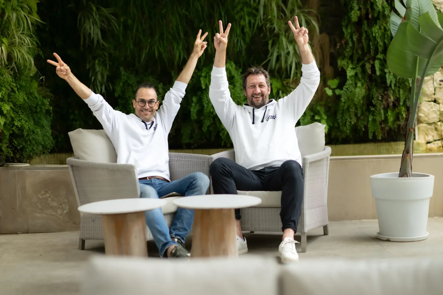 Two men in white hoodies with the EINO logo are sitting outside, both joyfully raising their arms in the air