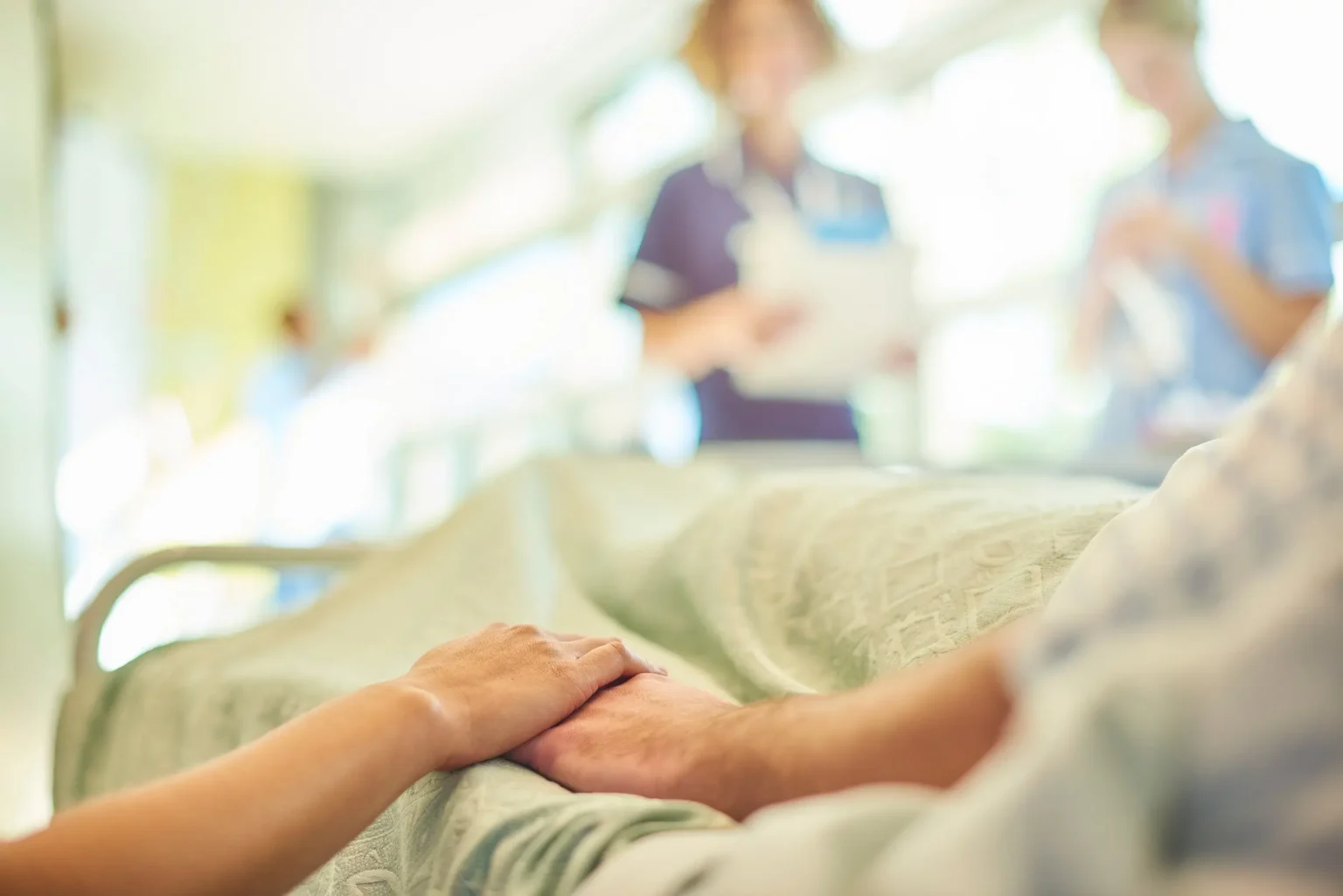 Hand holding another at the bedside, nursing staff out of focus in the background